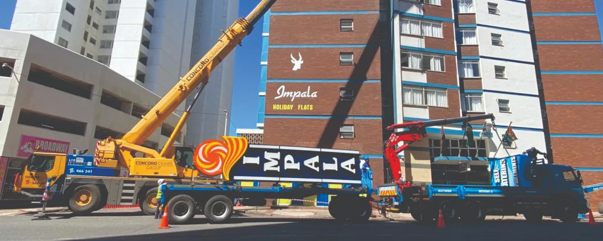 Large IMPALA building sign being lifted into place by crane outside Impala Holiday Flats